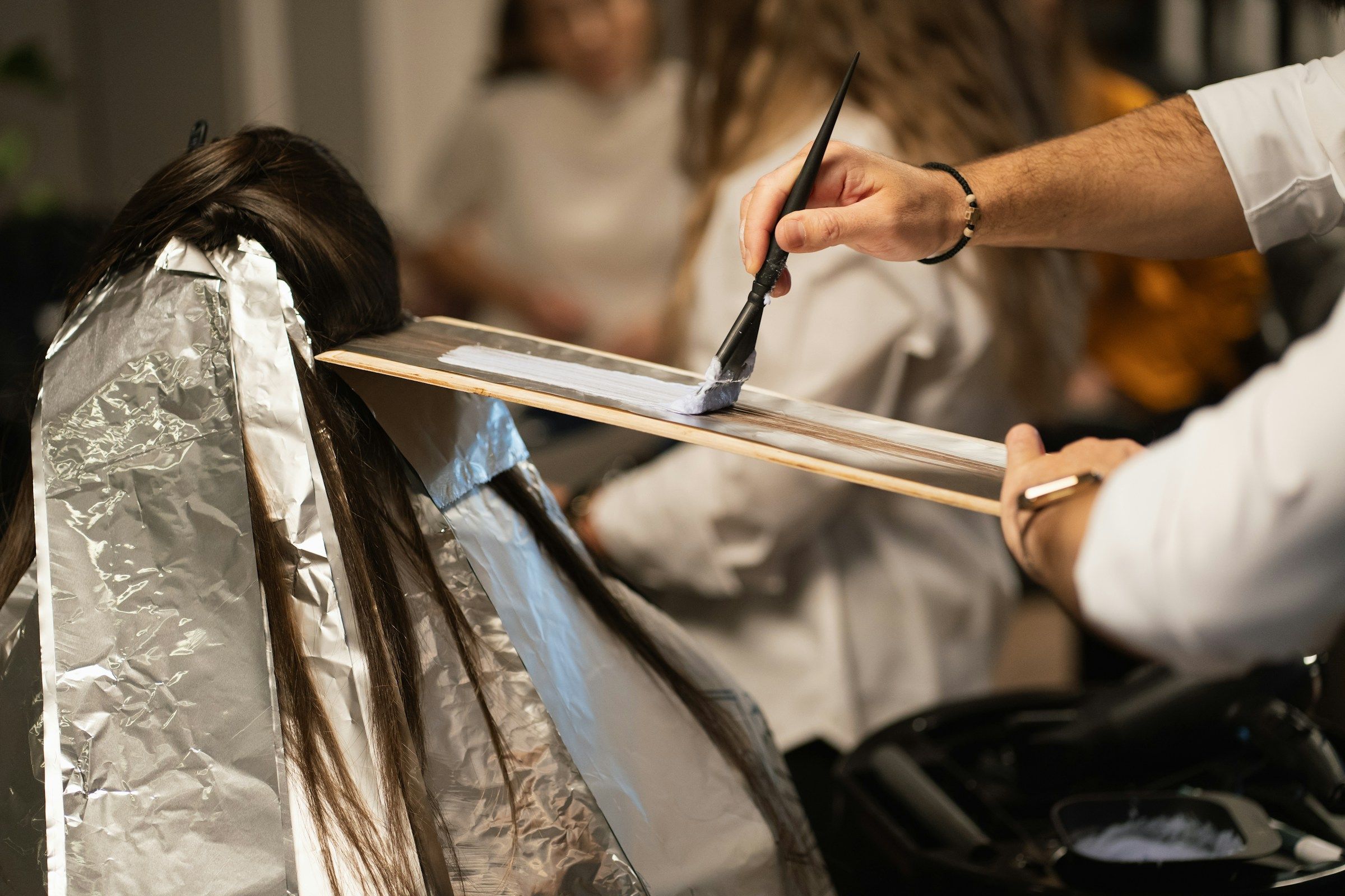 A colorist paints lightener onto a section of brown hair laid over a board, with foils already in