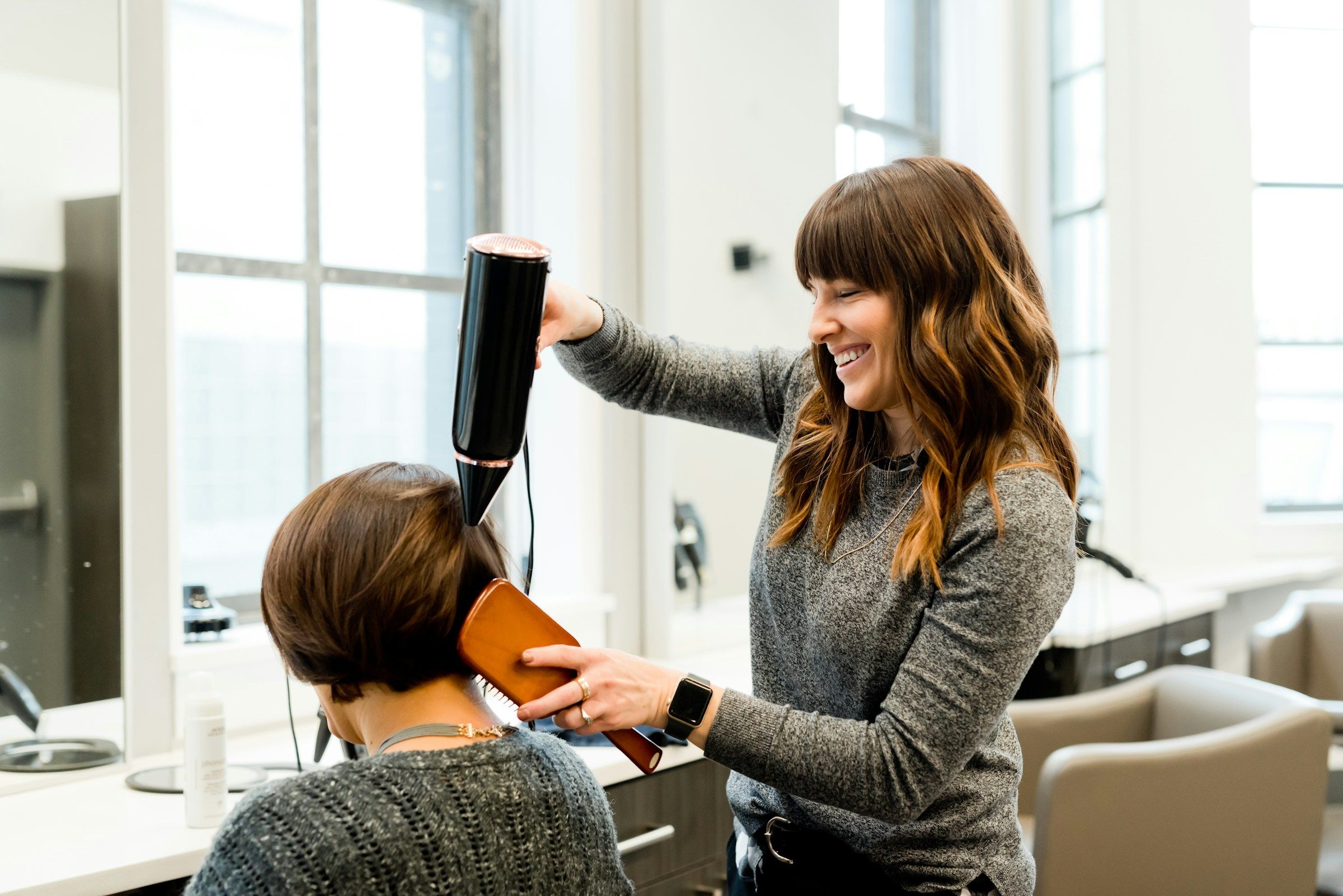 A stylist laughs as she blow-dries a client’s brown bob in a bright studio with tall windows