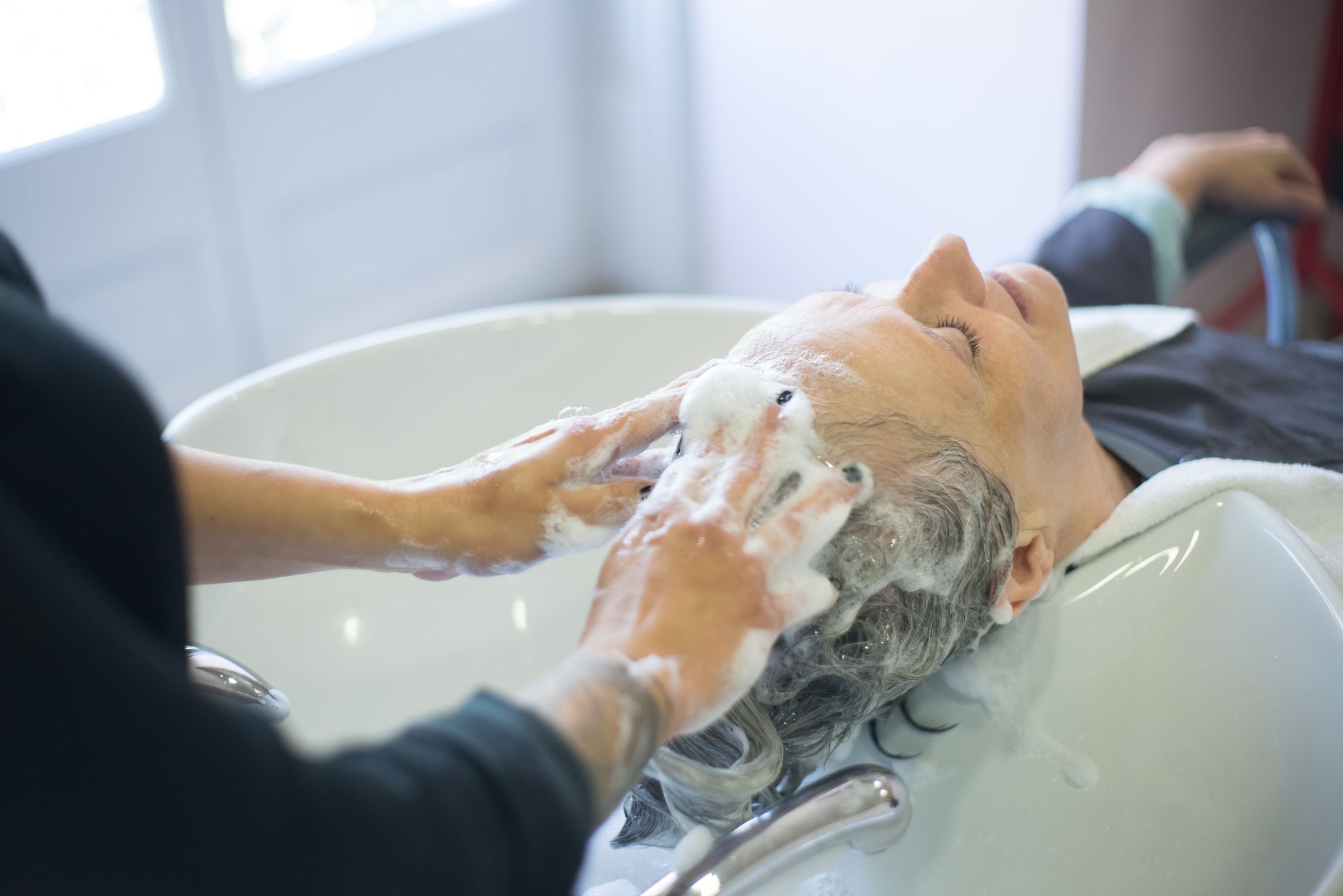 Shampoo being worked into an older woman’s gray hair at a white wash basin